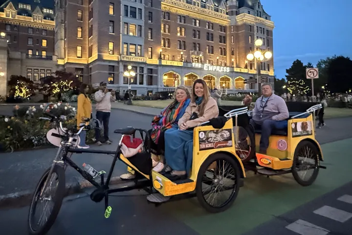 Three people in a pedicab in front of a lit-up historic building at dusk.